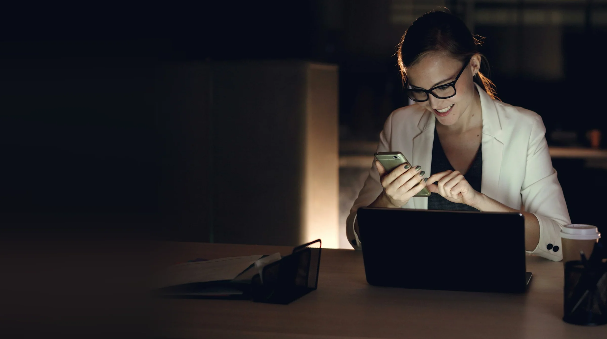 Woman at a laptop late in the evening, checking a text on her phone